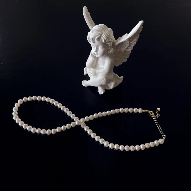 6mm imitation pearl necklace shaped as '8' in front of a white plaster angel, with a black background.