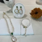 Freshwater pearl necklace, bracelet with green zircon accents, and drop earrings displayed on a white plaster base with a plaster angel and dry sunflower in the background.