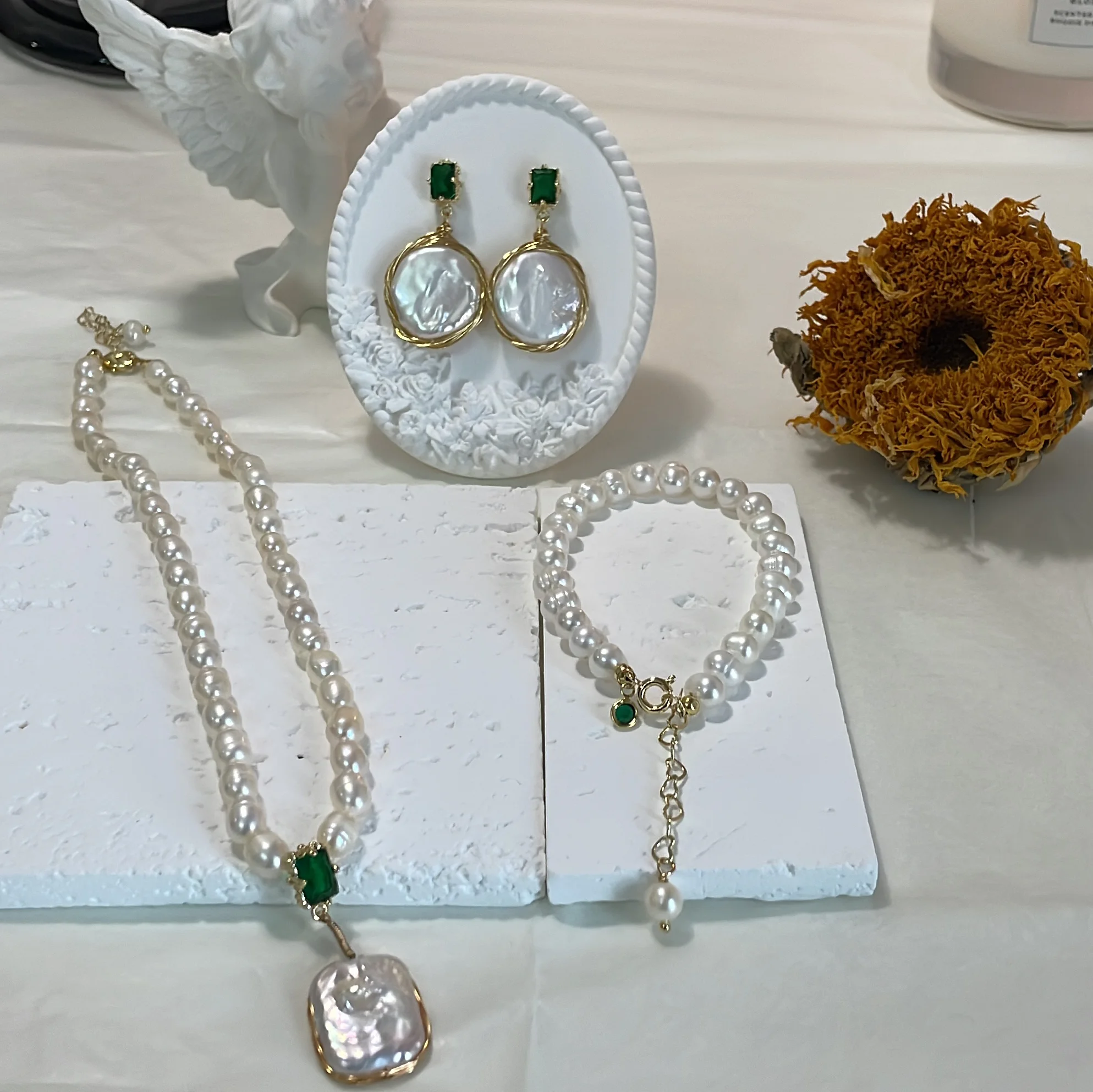 Freshwater pearl necklace, bracelet with green zircon accents, and drop earrings displayed on a white plaster base with a plaster angel and dry sunflower in the background.