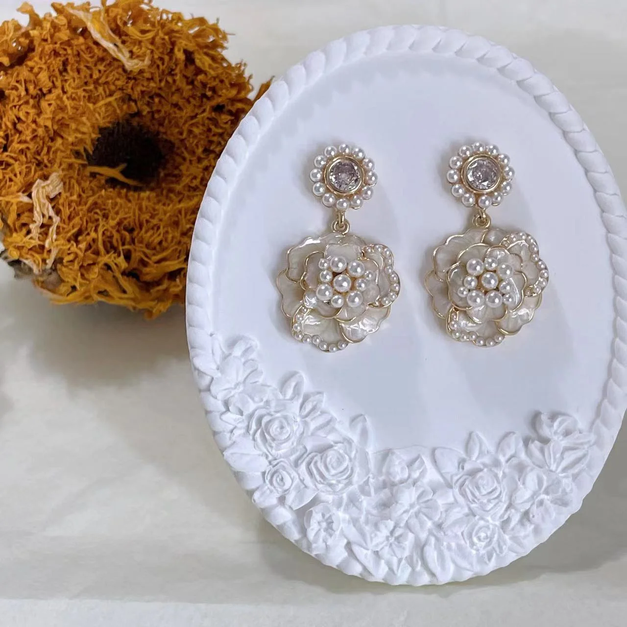Vintage camellia pearl drop earrings with enamel and rhinestones on white plaster stand with a dried sunflower behind.