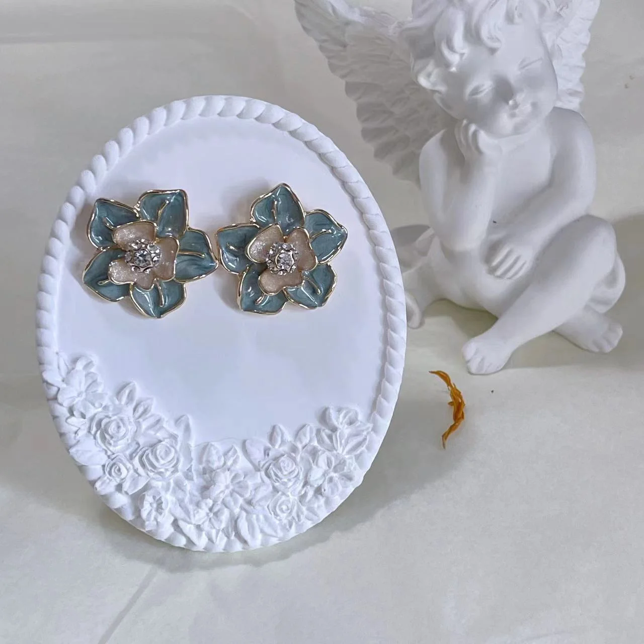 Vintage blue enamel camellia 14K gold-plated stud earrings on a white plaster stand with an angel statue in the background.