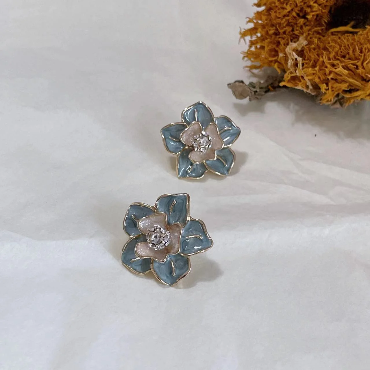 Vintage blue enamel camellia 14K gold-plated stud earrings placed on white fabric with a dried sunflower behind.