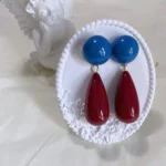 Vintage 14K gold-plated red and blue color block geometric earrings displayed on a plaster stand with an angel statue behind.